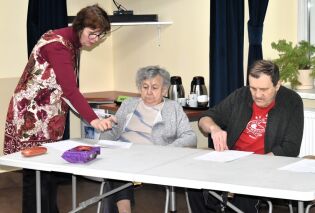 Kobieta pochyla się nad stołem i wskazuje dokumenty dwóm siedzącym osobom, które czytają kartki; scena rozgrywa się w jasnym pomieszczeniu z zasłonami i sprzętem do serwowania napojów w tle.