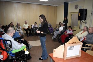 W sali terapeutycznej kilkanaście osób siedzi w kręgu, część na wózkach inwalidzkich. Prowadząca stoi pośrodku i trzyma element przedstawienia. Na pierwszym planie znajduje się drewniana skrzynka