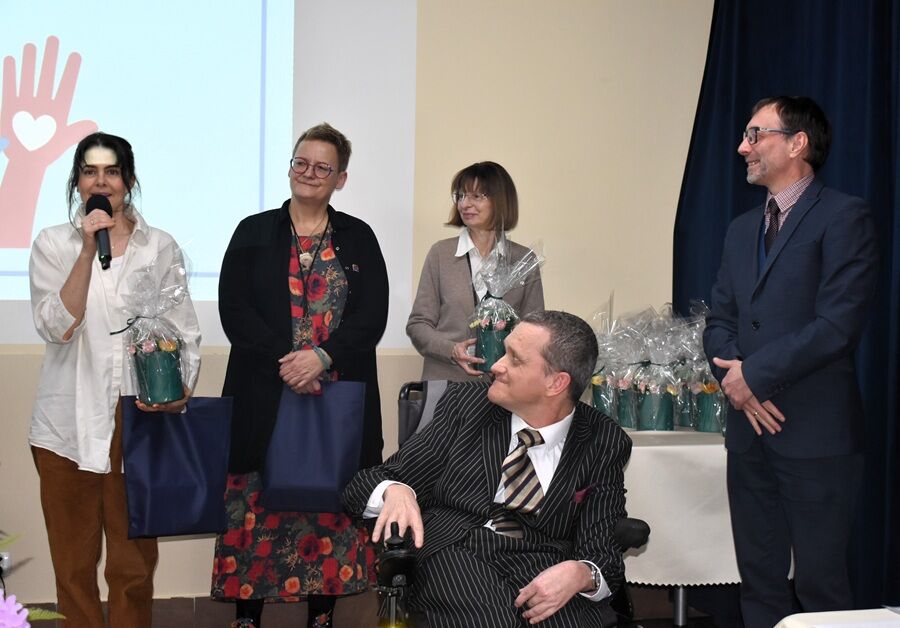 Cztery osoby stoją na scenie podczas uroczystości. Trzy kobiety trzymają upominki zapakowane w celofan oraz torby prezentowe, jedna z nich przemawia do mikrofonu. Obok nich stoi mężczyzna.