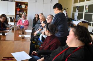 Grupa dorosłych osób, w tym seniorzy i osoby z niepełnosprawnościami, siedzi przy stole w jasnej sali. Na stole leżą kartki i długopisy. Kobieta stojąca obok jednego z uczestników podaje mu mikrofon. 