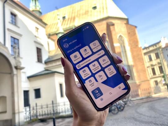 Ręka trzymająca telefon komórkowy z otwartą aplikacją mKraków. W tle widoczny kościół Franciszkanów przy ulicy Franciszkańskiej w Krakowie.