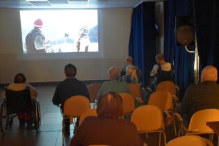 Grupa osób siedzi na krzesłach i ogląda film wyświetlany na dużym ekranie w sali przypominającej małe kino. Niektórzy widzowie korzystają z wózków inwalidzkich.