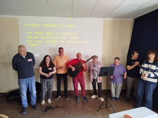 Grupa dorosłych osób stoi na scenie podczas zajęć teatralnych. Jedna z osób gra na gitarze, a pozostali uczestnicy stoją obok i biorą udział w występie. Na ścianie za nimi wyświetlona jest prezentacja