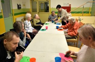 Kilka starszych osób siedzi przy stole w jasnym pomieszczeniu i uczestniczy w zajęciach sensorycznych. Każda osoba ma przed sobą talerz z kawałkiem ciasta i kolorowy kubek. 