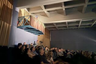 Publiczność ogląda spektakl w sali teatralnej, a nad widownią na scenicznym balkonie stoi kobieta. 