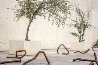 Grafika przedstawia wystawę sztuki nowoczesnej - na podłodze leżą wygięte duże gwoździe, a na betonowych podestach umieszczono drzewa liściaste.