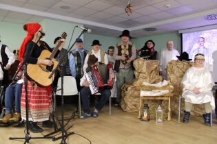 Scena jasełek z muzyką na żywo – gitarzystka i akordeonista oraz grupa aktorów zgromadzonych wokół symbolicznej stajenki.