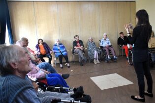 Grupa starszych osób siedzi w kręgu w sali, część z nich na wózkach inwalidzkich. Przed nimi stoi prowadząca, która pokazuje coś na rękach podczas zajęć muzykoterapii.