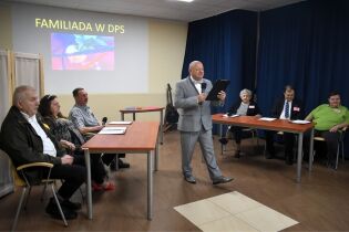 Mężczyzna w garniturze przemawia do siedzących uczestników podczas wydarzenia z okazji Dnia Kobiet. W tle widać prezentację na ekranie i instrumenty muzyczne.