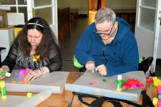 Dwie osoby siedzą przy stole i wykonują pracę plastyczną z kolorowego papieru, używając kleju. Na stole leżą materiały plastyczne i przygotowane elementy dekoracji.

