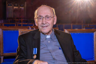 Ks. Prof. Michał Heller siedzi na krześle w auli Collegium Novum Uniwersytetu Jagiellońskiego. Na jego piersi widać odznakę Honoris Gratia.