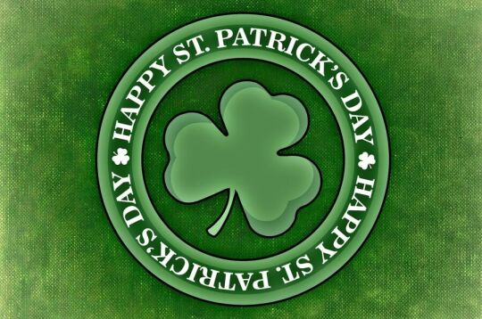 Grafika na której na zielonym tle imitującym tkaninę jest okrągła przypinka z trójlistną koniczyną w środku a w otoku napis Happy St. Patrick's Day