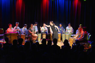 Widok z tyłu widowni: sylwetki osób siedzących na krzesłach, przed nimi scena. Na pierwszym planie dziewczynka w czarnym stroju, za nią kilkanaście osób grających na bębnach.