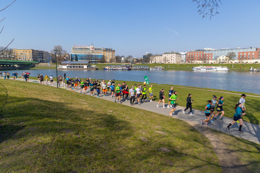 Biegacze z numerami startowymi biegną bulwarami wiślanymi w słoneczny dzień. Trasa prowadzi wzdłuż rzeki Wisły, w tle widoczna woda, zieleń oraz zabudowania. Uczestnicy poruszają się zwartą grupą.