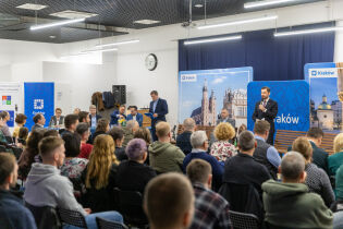 Spotkanie w sali: na pierwszym planie uczestnicy siedzą na krzesłach, w tle widoczni prelegenci, a za nimi ustawione banery informacyjne. Wydarzenie ma charakter otwartego spotkania i dyskusji.