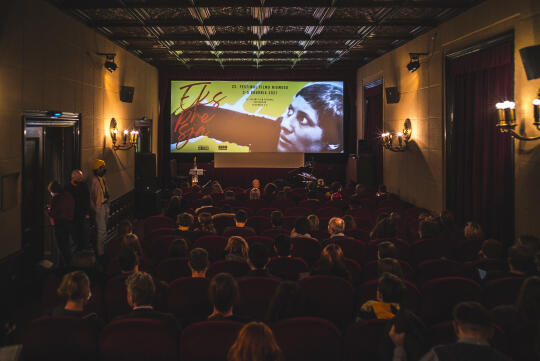 Kameralna, przyciemniona sala kinowa. Na pierwszym planie widzowie siedzą w fotelach, zwróceni w stronę ekranu. W tle widoczny jest ekran kinowy z wyświetlanym kadrem filmu.