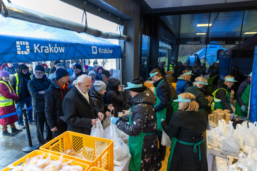 Lada przed budynkiem z zapakowanym żurkiem i pieczywem w żółtych koszach, ludzie wydają potrawę, a przed ladą stoi kolejka oczekujących pod namiotem z napisem ‚Kraków’.
