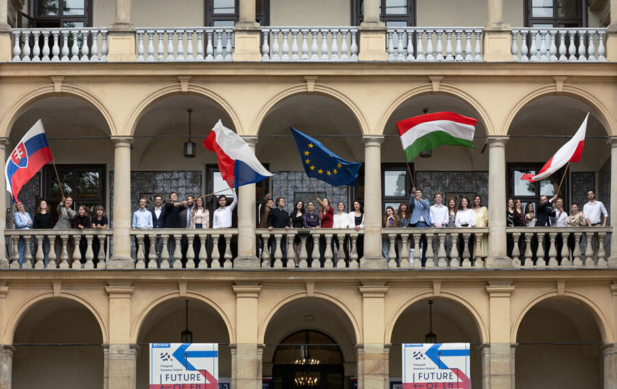 Budynek Willi Decjusza w którym na galerii stoi młodzież z krajów Grupy Wyszehradzkiej. cztery osoby trzymają za drzewce duże flagi Słowacji, Węgier, Czech i Unii Europejskiej. 