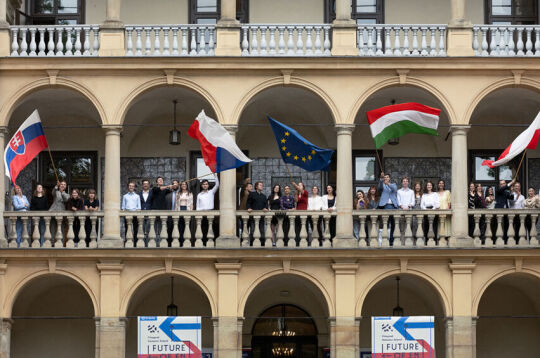 Budynek Willi Decjusza w którym na galerii stoi młodzież z krajów Grupy Wyszehradzkiej. cztery osoby trzymają za drzewce duże flagi Słowacji, Węgier, Czech i Unii Europejskiej. 