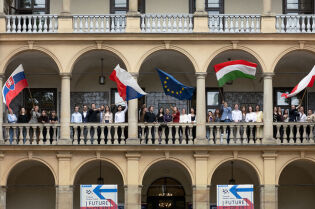 Budynek Willi Decjusza w którym na galerii stoi młodzież z krajów Grupy Wyszehradzkiej. cztery osoby trzymają za drzewce duże flagi Słowacji, Węgier, Czech i Unii Europejskiej. 