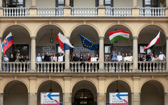 Budynek Willi Decjusza w którym na galerii stoi młodzież z krajów Grupy Wyszehradzkiej. cztery osoby trzymają za drzewce duże flagi Słowacji, Węgier, Czech i Unii Europejskiej. 