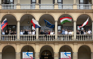 Budynek Willi Decjusza w którym na galerii stoi młodzież z krajów Grupy Wyszehradzkiej. cztery osoby trzymają za drzewce duże flagi Słowacji, Węgier, Czech i Unii Europejskiej. 