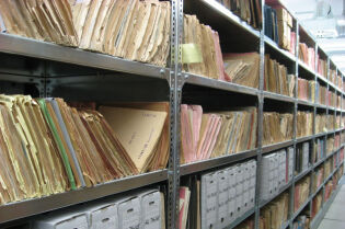 Grafika przedstawia stare dokumenty, ułożone na półkach.