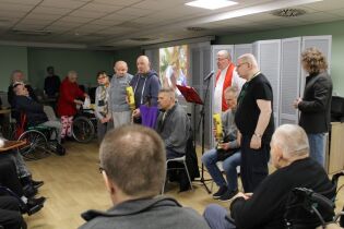 Grupa kobiet i mężczyzn, w tym osoby na wózkach inwalidzkich, uczestniczy w nabożeństwie. Duchowny prowadzi modlitwę przy mikrofonie, a część uczestników trzyma zapalone świece.