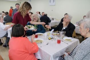 Kobieta podaje talerz z jedzeniem osobom siedzącym przy stole podczas wspólnego obiadu w jadalni.