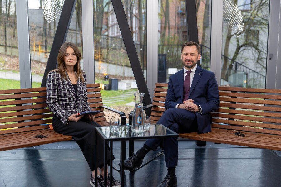 Pomieszczenie z dużymi oknami. Na ławkach siedzi prezenterka i PMK Aleksander Miszalski. Przed nimi mały stolik z dwoma szklankami i karafką wody. Za oknami trawa, drzewa i zabudowania.