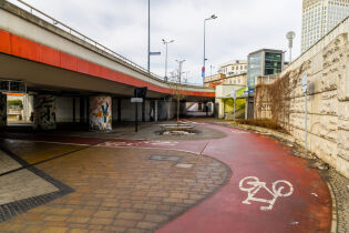 ścieżka rowerowa Rondo Mogilskie dolny poziom. Widoczna pusta czerwona wstęga ścieżki dla rowerów z oznaczeniem białego symbolu roweru. Widoczna estakada oraz budynki w tle.