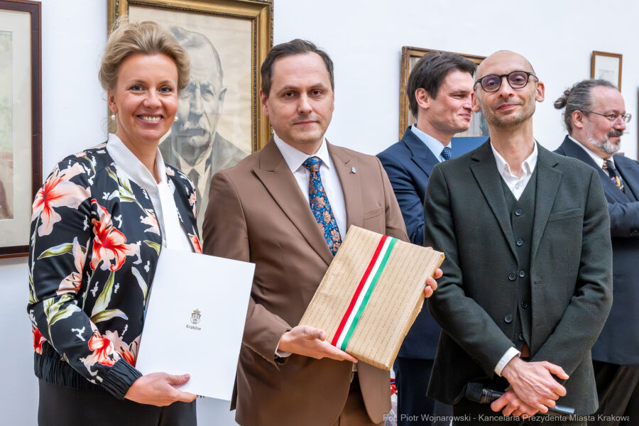 przed grafikami na ścianie wnętrza wystawienniczego stoja trzy osoby, od lewej jasnowłosa kobieta z upiętymi włosami, w środku Przewodniczący Rady Miasta Krakowa a po lewej Pełnomocnik Prezydenta 