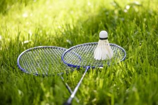 Grafika przedstawia dwie paletki do badmintona i lotkę, leżące na zielonej trawie.