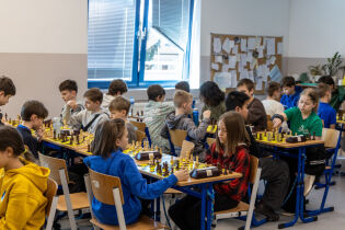 Na zdjęciu widać kilkanaścioro dzieci grających w szachy przy stołach ustawionych w rzędach w sali wewnątrz budynku. Uczestnicy siedzą naprzeciwko siebie, skupieni na partiach. Na stołach znajdują się