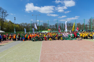 Na zdjęciu widać boisko piłkarskie oraz liczne dzieci w klubowych koszulkach podczas turnieju Futbol Plus. Na trybunach i przy boisku obecni są rodzice, którzy wspierają zawodników w trakcie sportowej