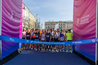 Uczestnicy TAURON 23. Cracovia Maraton na Rynku Głównym w Krakowie. Zawodnicy w strefie startu, z niebieską szarfą, czekają na rozpoczęcie biegu. Słoneczna pogoda i tłumy w centrum miasta.