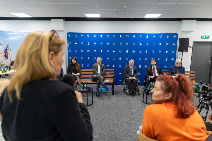 Pani z mikrofonem przemawia na pierwszym planie, obok siedzi druga kobieta. W tle przedstawiciele miasta Krakowa na ławkach oraz granatowo-niebieska ścianka z symbolami Krakowa. Sala z białym sufitem 