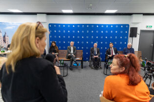 Pani z mikrofonem przemawia na pierwszym planie, obok siedzi druga kobieta. W tle przedstawiciele miasta Krakowa na ławkach oraz granatowo-niebieska ścianka z symbolami Krakowa. Sala z białym sufitem 