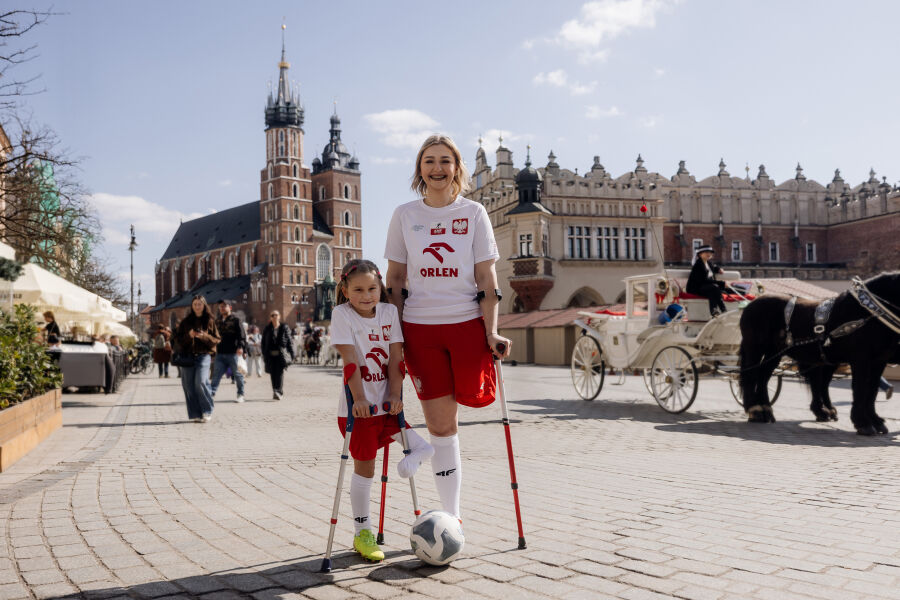 Uśmiechnięta jasnowłosa młoda kobieta po amputacji nogi w polskim narodowym stroju piłkarskim stoi o kulach na Rynku Głównym na tle. Przytula się do niej mała dziewczynka również po amputacji.   