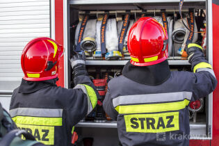 Przy otwartym z boku samochodzie pożarniczym w którym widać urządzenia gaśnicze stoi tyłem dwóch strażaków w czerwonych hełmach wyjmujących elementy gaśnicze. 