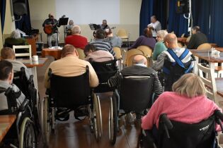 Osoby na wózkach inwalidzkich siedzą w sali i słuchają występu muzycznego. Na niewielkiej scenie kilku muzyków gra na gitarach i śpiewa, a publiczność jest zwrócona w ich stronę.