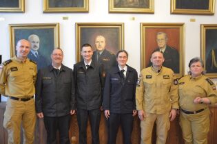Wizyta delegacji strażaków z Frankfurtu nad Menem w Krakowie. Pod portretami różnych ważnych w historii straży krakowskiej postaci stoi sześć uśmiechniętych osób w mundurach. Dwie z nich to kobiety.