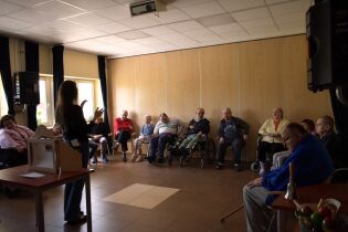 Grupa osób siedzi w kręgu w sali, uczestnicząc w zajęciach prowadzonych przez stojącą kobietę. Niektórzy uczestnicy korzystają z wózków inwalidzkich. Pomieszczenie jest przestronne, z jasnymi ścianami