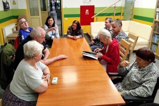 Grupa starszych osób siedzi przy dużym stole w przestrzeni wspólnej i bierze udział w spotkaniu biblioterapeutycznym. Jedna z uczestniczek trzyma otwartą książkę lub album, a pozostali słuchają.