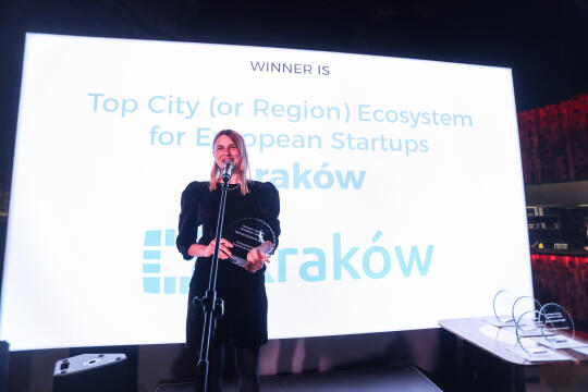 Elegancko ubrana kobieta trzyma w ręce szklana statuetkę w kształcie półkolistym. W tle ekran w napisem Top European City Ecosystem for Startups the winner is: Kraków.   