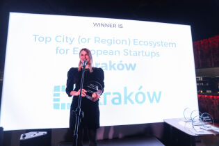 Elegancko ubrana kobieta trzyma w ręce szklana statuetkę w kształcie półkolistym. W tle ekran w napisem Top European City Ecosystem for Startups the winner is: Kraków.   