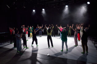Na ciemnej scenie grupa tancerzy wykonuje układ choreograficzny na białej płachcie. Środek sceny oświetla kilka reflektorów, podkreślając ruch i sylwetki artystów.