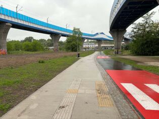 Pochmurny, deszczowy dzień. Obok ścieżki rowerowej i chodnika rozciąga się pas zieleni, a nad nimi widoczne są dwa przejazdy kolejowe. Mokra nawierzchnia i szare niebo podkreślają spokojny, jesienny 