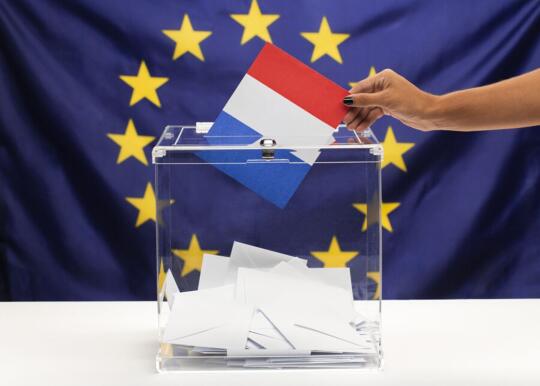 Urna Wyborcza na tle flagi Unii Europejskiej i kobieca ręka trzymająca symboliczna flagę Francji 