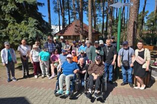 Grupa kobiet i mężczyzn pozuje do wspólnego zdjęcia w plenerze na tle wysokich drzew oraz drewnianej altanki znajdującej się na terenie ośrodka wypoczynkowego.